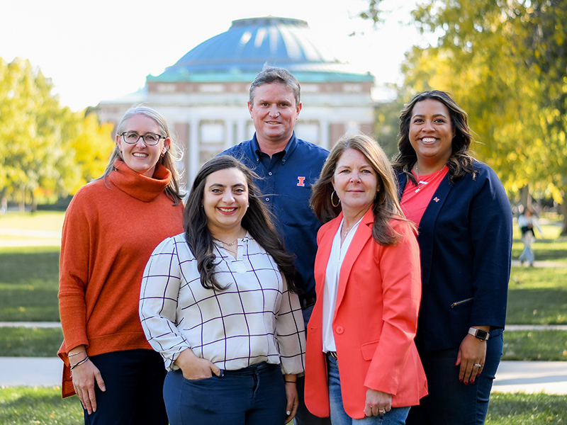 Five FSA professional staff members posing with Main Quad and Foellinger Auditorium in background